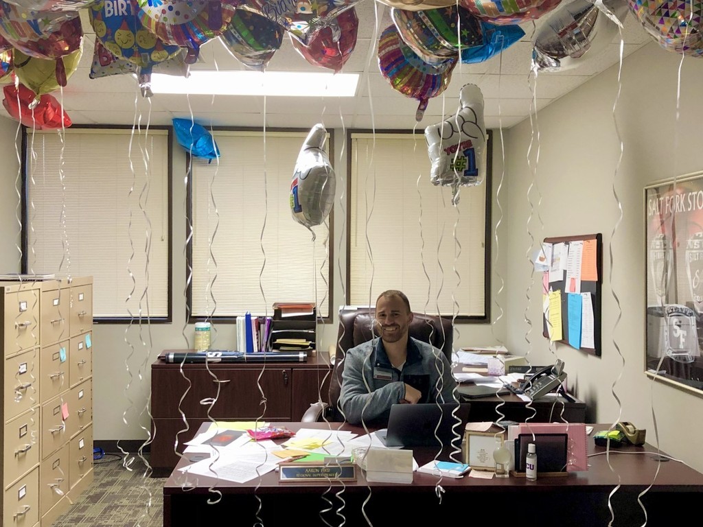 Aaron Hird sitting at his desk surrounded by balloons